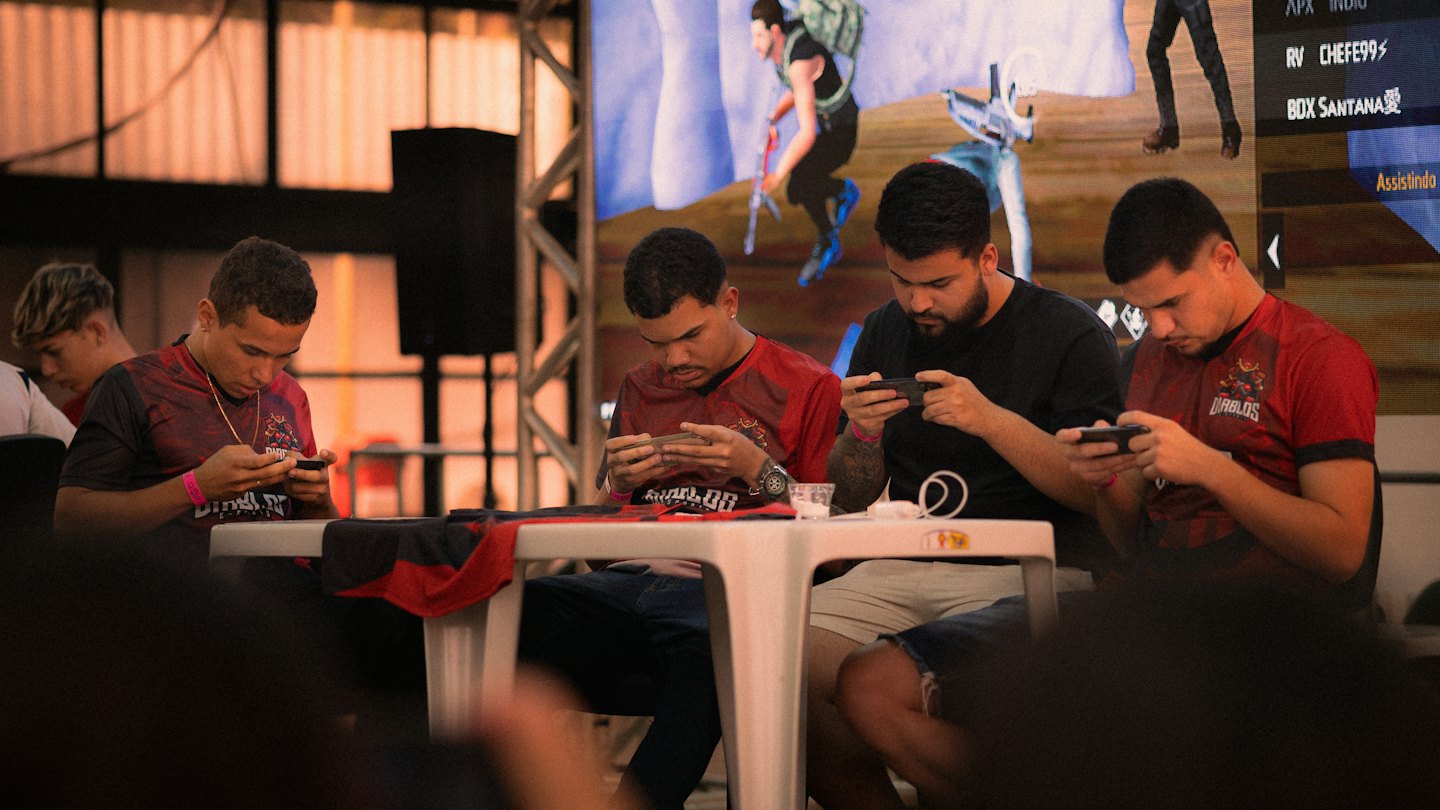 Four men playing mobile games on smartphones at a table.