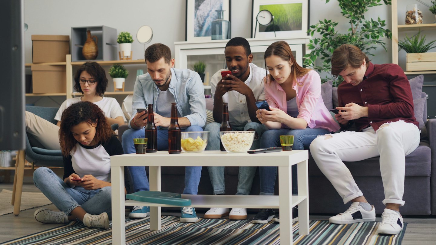 Six friends sit in a living room using their smartphones.
