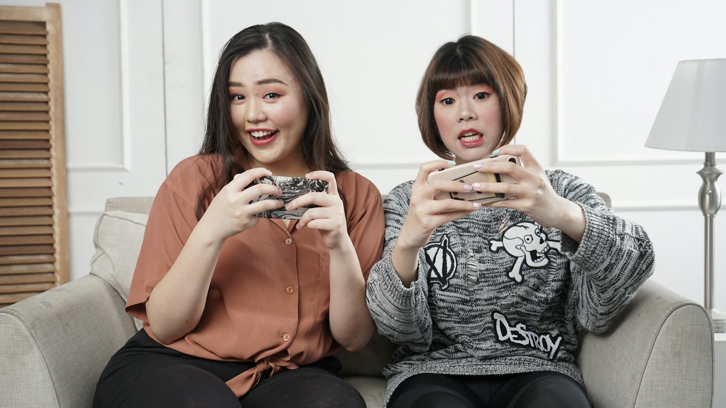 Two women play mobile games on a grey sofa.